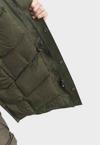 Doudoune vert olive avec sections rembourrées, une fermeture éclair et des boutons-pression. Tissu texturé mettant en valeur un intérieur lisse et isolé.