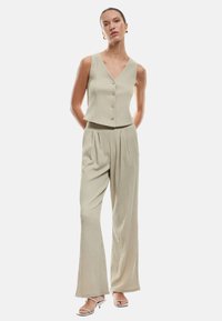 Vaaleanbeige lyhyt liivi, jossa on pystysuora rakenne ja kultaiset napit, yhdistetty leveälahkeisiin housuihin samasta kankaasta, jotka korostavat rentoa istuvuutta.