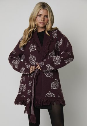 Blonde vrouw die een paarse gebreide cardigan met paisleypatroon en franjes over een zwart jurkje en zwarte panty sluit tegen een effen achtergrond.