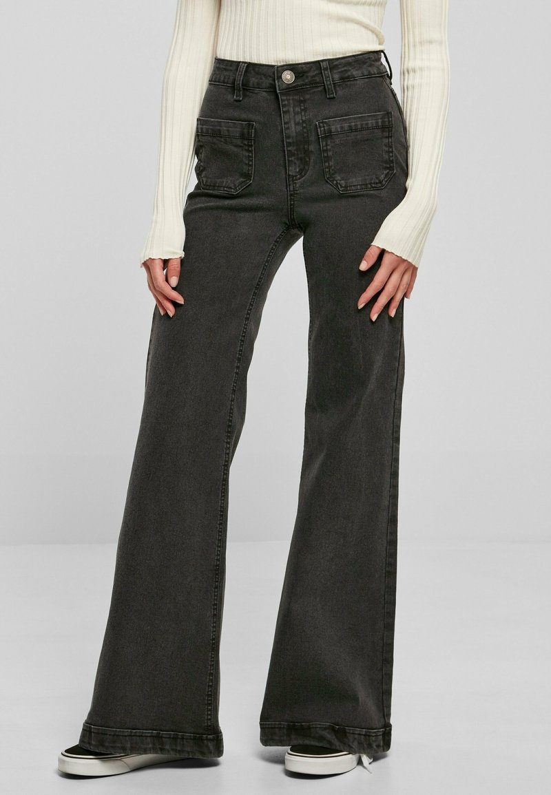 Femme portant un jean noir taille haute à jambes larges avec poches plaquées devant, associé à un haut blanc côtelé à manches longues et des baskets noires.