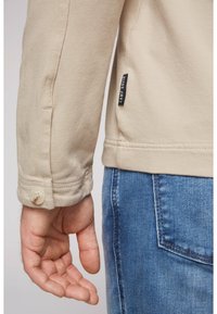Chaqueta beige con un tejido texturizado, que presenta un puño abotonado y una etiqueta en el costado. LLevada con jeans de mezclilla azules. Primer plano de la manga.