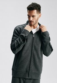 Sweter z zamkiem błyskawicznym w ciemnoszarym kolorze, wykonany z teksturowanej tkaniny, z prążkowanymi mankietami i akcentami logo na rękawach. Zamek z przodu.