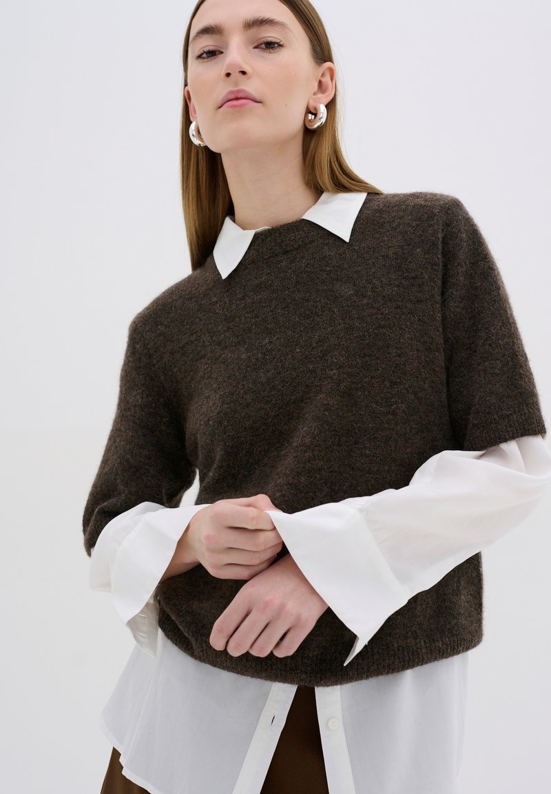 Pull en laine marron avec un col rond, superposé sur une chemise blanche à col, dotée de larges poignets. Texture lisse et coupe décontractée.