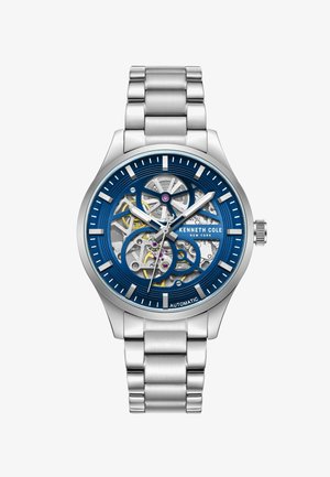 Automatische Uhr mit einem Armband aus Edelstahl. Das blaue Zifferblatt weist ein skelettiertes Design, weiße Stundenmarkierungen und ein sichtbares Uhrwerk mit goldenen Akzenten auf.