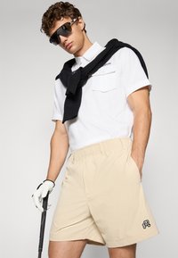 Beige shorts med elastisk midje och svart broderad logotyp, kombinerat med en vit pikétröja och en svart tröja över axlarna. Svarta solglasögon.