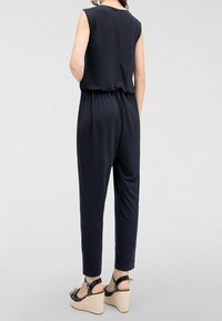 Navy jumpsuit med en figursydd överdel, elastisk midja och avsmalnande ben. Har korta ärmar och dragkedja i ryggen. Bärs med svarta kilklackar.