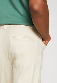 Osoba v béžových kalhotách a zeleném tričku, se pravou rukou v kapse kalhot, zobrazená od boků k polovině těla na jednobarevném pozadí.