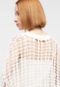 Λευκό πλεκτό μπολερό με σχέδιο ανοιχτού πλέγματος, υφή πλεξίματος και ριμπ λαιμόκοψη, φορεμένο πάνω από ανοιχτόχρωμη μπλούζα.