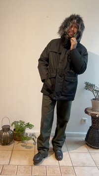 Parka preto com capuz de pelúcia, dois bolsos frontais com remendo, bolsos laterais e um fecho de correr. Calças verde-oliva e botas pretas. Ambiente interno.