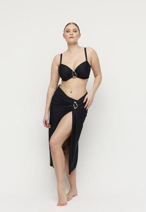 Vrouw poseert op blote voeten, draagt een zwarte bikinitop en een bijpassende sarongrok met hoge split en gouden gespdetails, op een witte achtergrond.