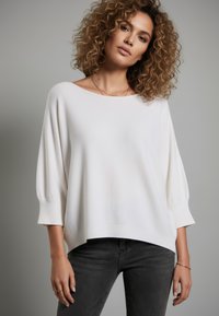 Weißer übergroßer Pullover mit weitem Ausschnitt und dreiviertellangen Ärmeln, aus weichem Strickstoff. Kombiniert mit dunkelgrauen Jeans.