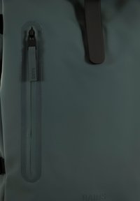 Mochila impermeable de color verde oscuro, con una textura suave, bolsillo lateral con cremallera y un elemento de marca, y herrajes en negro contrastante.