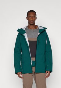 Muškarac u tirkiznoj vanjskoj jakni preko prugaste sive i smeđe majice, s bež hlačama, stoji naslonjen na jednostavnu sivu pozadinu.