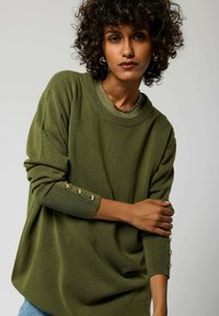 Maglione verde oliva con una vestibilità morbida, polsi a coste e dettagli in oro sui bottoni delle maniche. Tessuto a maglia morbido, scollatura rotonda.