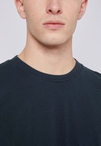 Tamsiai mėlynas medvilninis marškinėliai su klasikiniu apvaliu kaklu, trumpomis rankovėmis ir lygiu paviršiumi. Priartintas kaklo ir audinio detalių vaizdas.