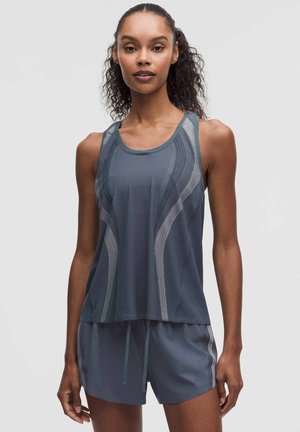 Mujer con cabello rizado lleva una camiseta deportiva sin mangas de color gris oscuro y pantalones cortos a juego sobre un fondo claro y liso.