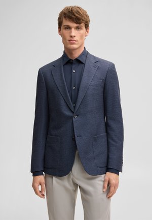 Jeune homme portant un blazer bleu marine sur une chemise bleu foncé boutonnée et un pantalon beige clair, debout devant un fond clair uni.