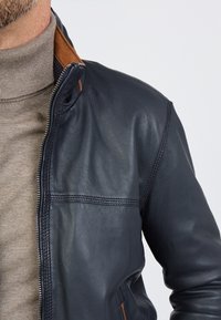 Marine leren jas met een ritsluiting en een bruine accenten bij de kraag. Kenmerkt zich door een gladde textuur en zichtbare stiksels langs de mouwen.