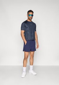 Tengerészkék sportpóló finom mintával, tengerészkék rövidnadrággal párosítva. A fehér tornacipő és a kék napszemüveg tökéletessé teszi az összhatást.