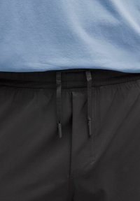 Detailný záber na čierny elastický pás s šnúrkami na nohaviciach v kombinácii s svetlomodrou košeľou.