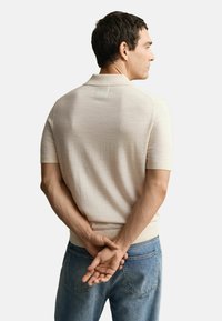 Man in beige short-sleeve knit polo shirt with collar stands facing away, hands clasped behind back, wearing blue jeans.