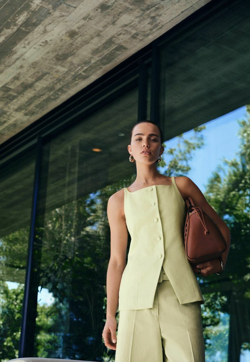 Donna che indossa una maglia gialla chiara senza maniche e pantaloni, con una borsa marrone in mano, in piedi davanti a una grande finestra di vetro che riflette gli alberi.