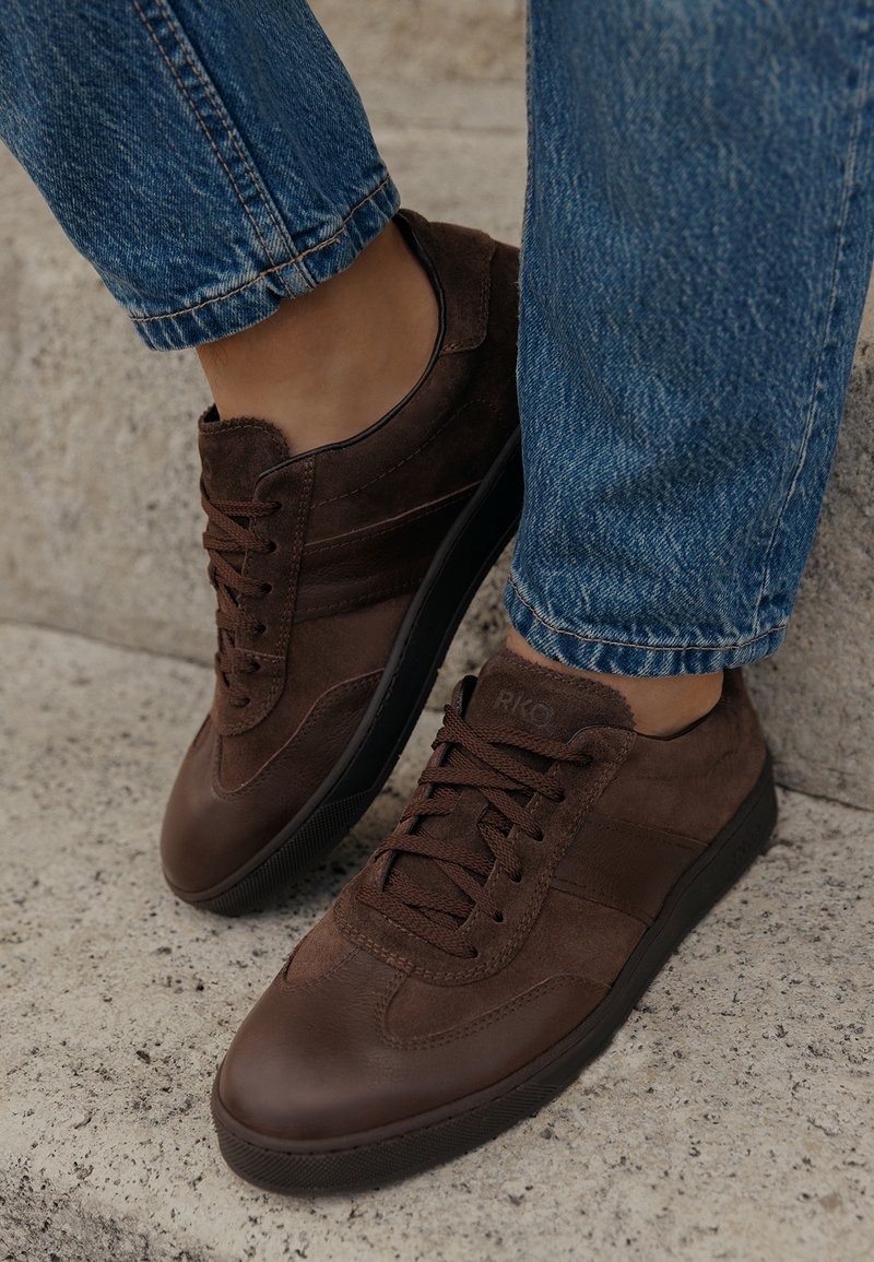Sneakers marroni in pelle e suede indossati con jeans blu su una superficie di pietra testurizzata, che mostrano i dettagli delle calzature e i lacci.