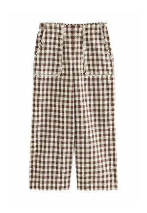 Frau steht auf dem Deck eines Segelbootes, trägt einen ärmellosen karierten Overall und braune Sandalen, mit dem Ozean und klarem Himmel im Hintergrund.