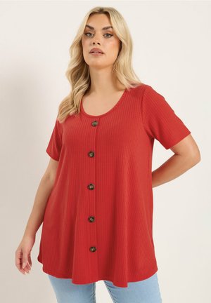 Kvinde med langt blondt hår iført en løs rød ribbet kortærmet top med store knapper foran og lyseblå jeans.