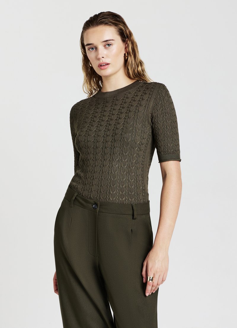 Haut tricoté à manches courtes couleur olive avec motif en câble texturé, associé à un pantalon taille haute assorti et une bague en argent au doigt.