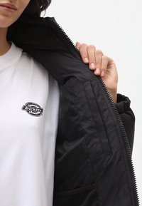 Czarna kurtka puchowa o gładkiej fakturze, zapinana na zamek, z wewnętrzną kieszenią; noszona na białym t-shircie z małym logo.