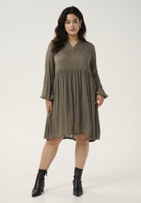 Robe vert olive à manches longues, décolleté en V et taille froncée. Présente des pois noirs et arrive à mi-mollet. Portée avec des bottines noires.