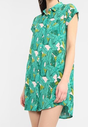 Vrouw in een korte mouw, jurk met knopen en een groen tropisch bladerenpatroon met een kleine kraag, staand tegen een effen achtergrond.