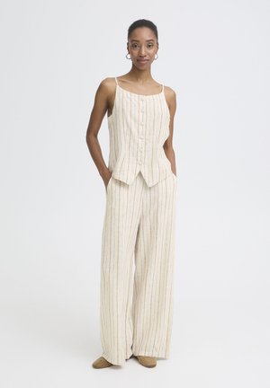 Donna che indossa un top beige senza maniche con bottoni e pantaloni abbinati a gamba larga con sottili righe verticali, in piedi con le mani nelle tasche.