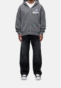 Sudadera gris con cremallera, bordado de mariposa y bolsillos delanteros, combinada con pantalones negros de corte suelto y zapatillas en blanco y negro.