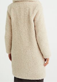 Abrigo de teddy en color beige con una textura esponjosa, mangas largas y cuello de chal. Tela suave y cálida, que llega hasta la mitad del muslo.