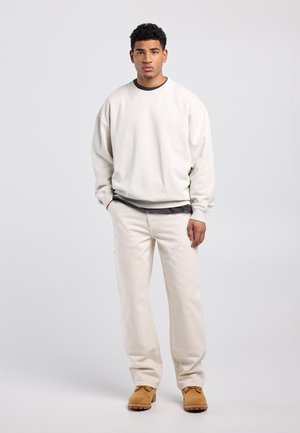 Junger Mann steht mit den Händen in den Taschen, trägt ein beigefarbenes Sweatshirt, passende Hose und beige Schnürstiefel vor einem schlichten grauen Hintergrund.