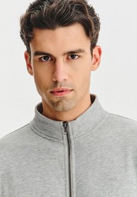 Szara kurtka zapinana na zamek z kołnierzem typu stójka i gładką fakturą. Posiada przedni zamek błyskawiczny oraz subtelne logo na wysokości klatki piersiowej.