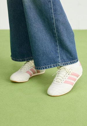 Witte sneakers met roze strepen gedragen met wijde blauwe spijkerbroeken op een groene vloer.
