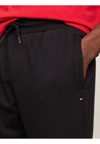 Svarta sweatpants med dragsko i midjan, sidofickor och en liten logotyp i röd, vit och blå. Mjuk tygtextur.