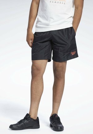 Person iført sorte Reebok-shorts, en hvid skjorte og sorte sneakers, står med hænderne i lommerne på en lys baggrund.