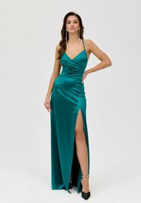 Tyrkysová saténová večerní róba s hlubokým výstřihem, nařaseným pasem a vysokým rozparkem, doplněná černými podpatky a stříbrnými náušnicemi.