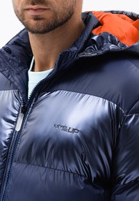 Marine pufferjack met een glanzende afwerking, oranje binnenvoering, logo op de borst, ritssluiting en hoge kraag. Getextureerde stof, geïsoleerd ontwerp.