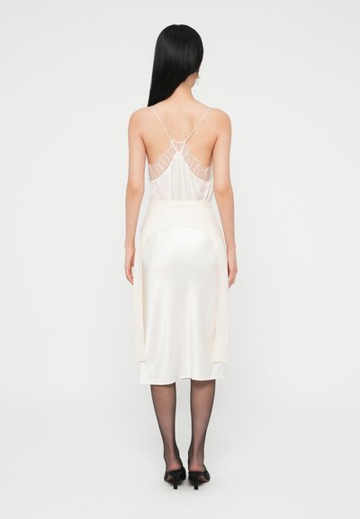 Robe en satin crème avec un dos décolleté et de fines bretelles, ornée d'une délicate dentelle. Texture lisse, longueur genou, avec des accents de drapé sur les côtés.