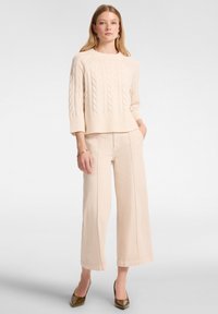 Maglione beige a maglia grosso con orlo a coste, abbinato a pantaloni larghi beige chiaro. Le scarpe hanno la punta appuntita di un verde scuro.