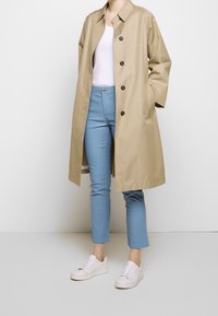 Impermeabile beige con chiusura ai bottoni, indossato sopra una canotta bianca, abbinato a pantaloni slim blu e sneakers bianche. Tessuto liscio.