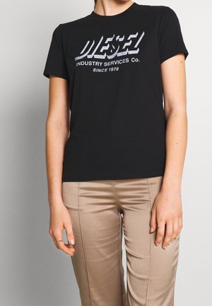 Kvinde iført en sort Diesel T-shirt med korte ærmer med hvid tekst og beige bukser, står mod en ensfarvet lysegrå baggrund.