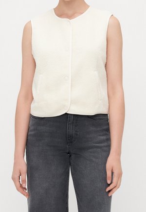 Gilet blanc sans manches texturé à boutons porté avec un jean noir délavé taille haute sur une personne devant un fond uni.