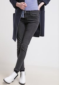 Svarta skinny jeans med en figurnära design och subtil stretch, kombinerade med vita ankelstövlar och en randig skjorta under en marinblå kappa.