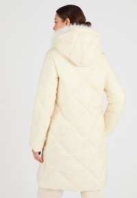 Lunga giacca trapuntata di colore crema, con cappuccio foderato in pelliccia, delicato motivo a rombi e una vestibilità rilassata con maniche lunghe.